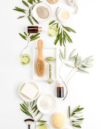 Flat lay of natural skincare products, including bottles, a brush, jade roller, soap, sponges, bowls of powders, and green leaves arranged on a white background.