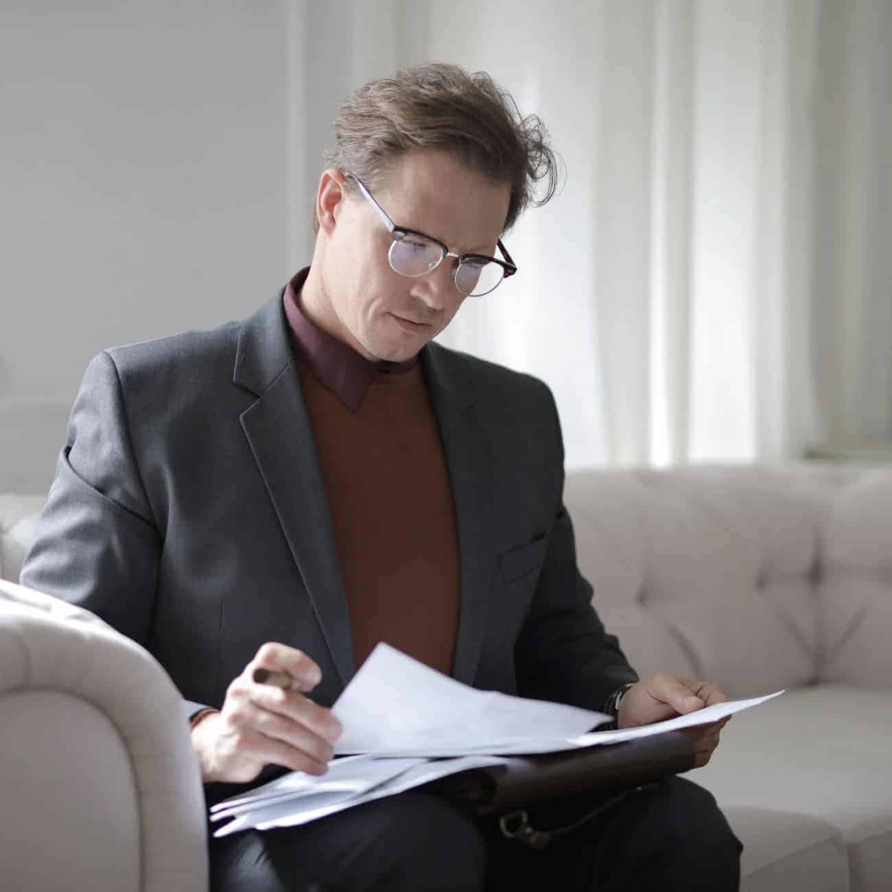 Man in glasses sitting on a couch, wearing a suit and reviewing documents.