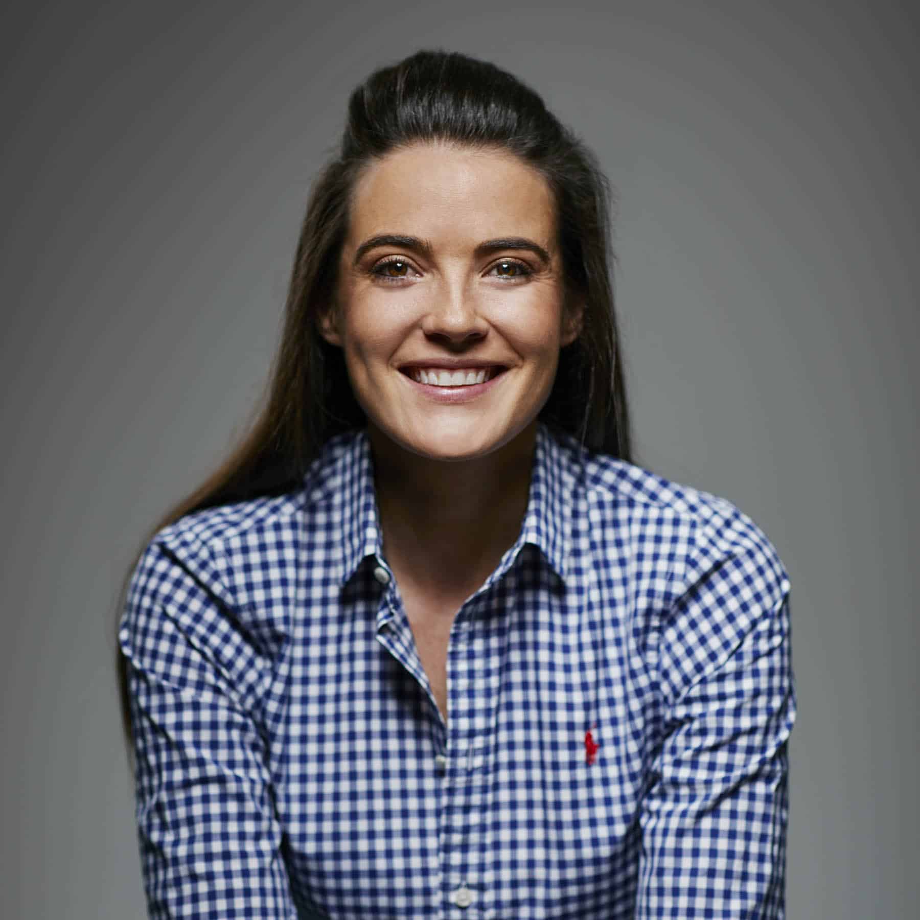 Person smiling, wearing a blue and white checkered shirt, with long hair and a neutral background.