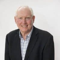 An older man with short white hair smiles while wearing a dark blazer over a checkered shirt, against a plain background.